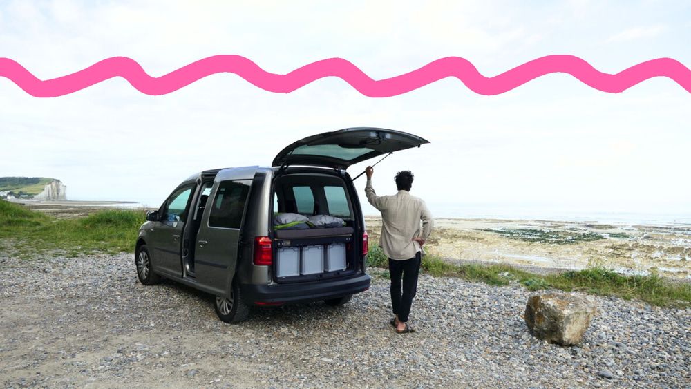 Stefan steht neben der offenen Heckklappe eines VW Caddys an einer Meeresküste.