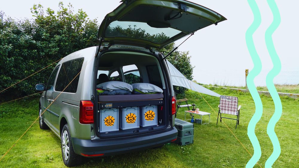 Ein Minicamper steht auf einem Campingplatz am Meer. Vor dem Minicamper stehen zwei Stühle, ein Tisch und ein Sonnensegel.