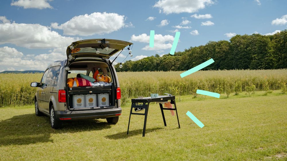 Ein Caddy mit Campingbett und mobiler Campingküche steht auf einer grünen Wiese. 